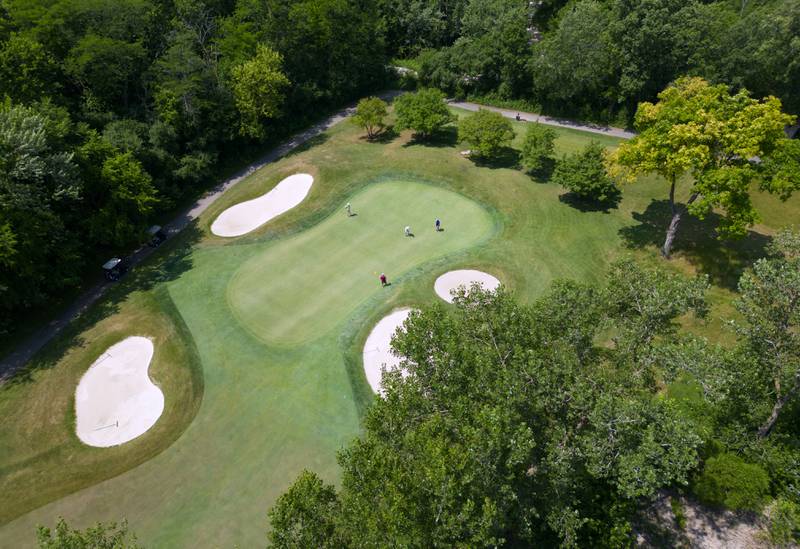 Golfers putt on the green on hole 5 west at Arrowhead Golf Club in Wheaton. The green is surrounded by trees and a natural wetland, bottom right.