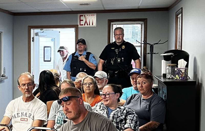 Waterman Police Chief Jason Swanson, standing in the back left of a crowded Waterman Village Trustee Board meeting on July 9, 2024, joined a cacophony of public comments from Waterman residents seeking to understand why he was suspended without pay earlier in the year.