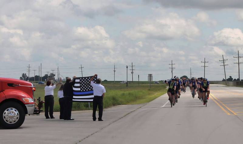 Law enforcement including first responders from Waterman Fire Department hold a flag honoring police to greet nearly 100 cyclists biking from La Salle County to DeKalb on Saturday, July 12, 2025, for Cycle Across Illinois, a charity campaign to honor fallen police officers and raise funds for their families.