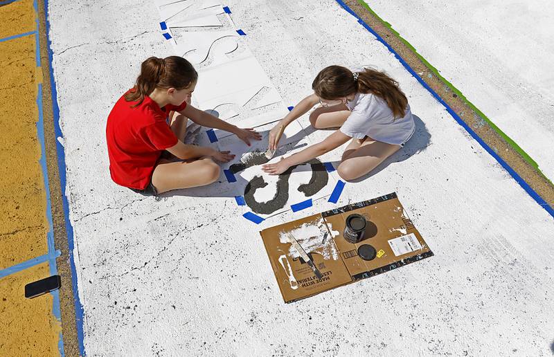 Marian Central Catholic High School  sophomore Abigail Huff helps her senior  sister, Juliette Huff by holding down the stencil that Juliette is using as she paints her parking spot on Tuesday, Aug. 13, 2024, before they to return to school on Thursday, Aug. 15, at the Woodstock school. This is the third year that the school's students have been able to bid at an auction to paint their parking spot.