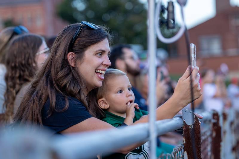 (from left) Katie Mcmillin holds son Benji as she records Boppin' Bruins performance before game on Friday, Sept 12, 2025 at Captain Thomas J. Heitmann field in Peru.