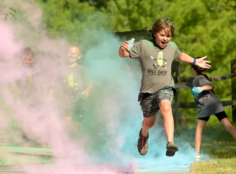 A pickle costumed Dill Dash runner leaps through cloud of colorful powder along the race route during the Inaugural Pickle Paradise event at Pottawatomie Park in St. Charles on Friday, June 21, 2024.