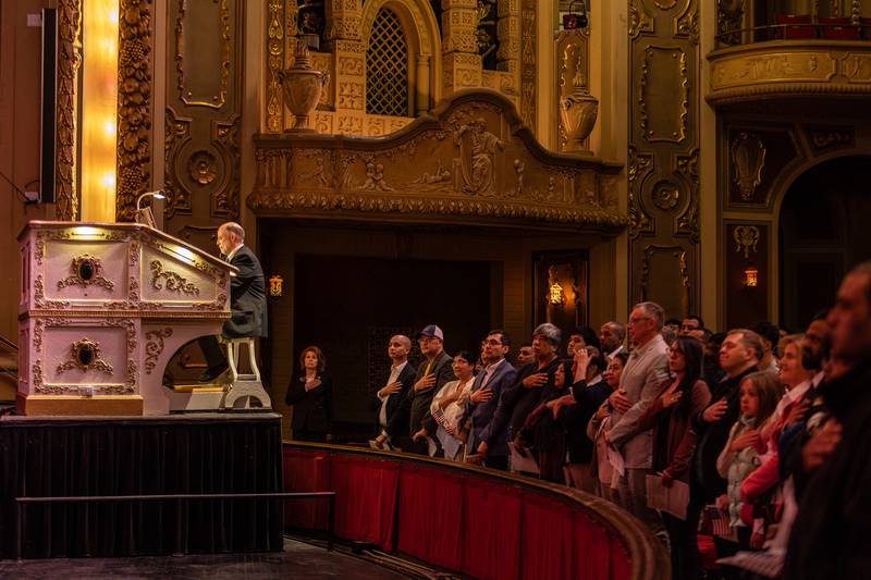 New U.S. citizens and attendees stand for the National Anthem performed by Rialto Square Theatre organist Taylor Trimby during the Naturalization Ceremony in Joliet on April 17, 2025.