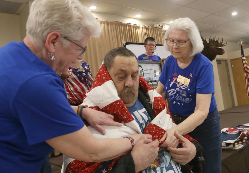 Veteran Gregory Zerfas, clenches a Quilt of Valor as Betty Baznik (left) and Shirley Steele (right) drape the quilt over him during the Quilt of Valor ceremony on Saturday, April 19, 2025 at the Moose Lodge in Princeton.  In 2003, quilter Catherine Roberts started the movement that became Quilts of Valor when her son was deployed in Iraq. Since that time, more than 401,361 have been awarded. The local group Illinois Valley Quilts of Valor awarded 189 quilts in 2024 and has given more than 1,000 quilts since its start in April 2017. Two veterans were absent.