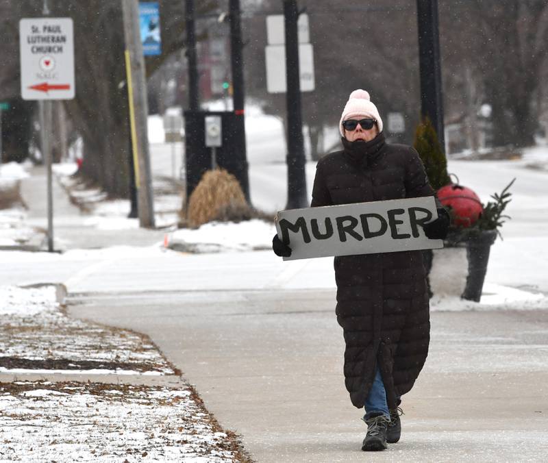 A woman carries the sign "murder" as she walks toward Indivisible of Ogle County's protest against ICE held Sunday, Jan. 25, 2026 in downtown Oregon. Approximately 120 people attended the afternoon event despite frigid winter temperatures to protest the shooting death of Alex Pretti by ICE agents on Saturday in Minneapolis.