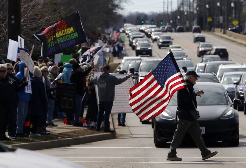 A man carries as American Flag as protesters line State Route 31 near the intersection of McCullom Lake Road in McHenry to protest their discontent with President Donald Trump and his administration's policies on Saturday, March 28, 2026, during the McHenry County No Kings Protest. According to an organizer, over 4,000, people took part in the protest.