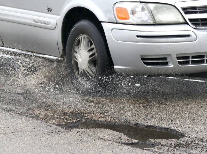 A vehicle splashes through a pothole Thursday on the entry road to Market Square Shopping Center from Sycamore Road in DeKalb.
