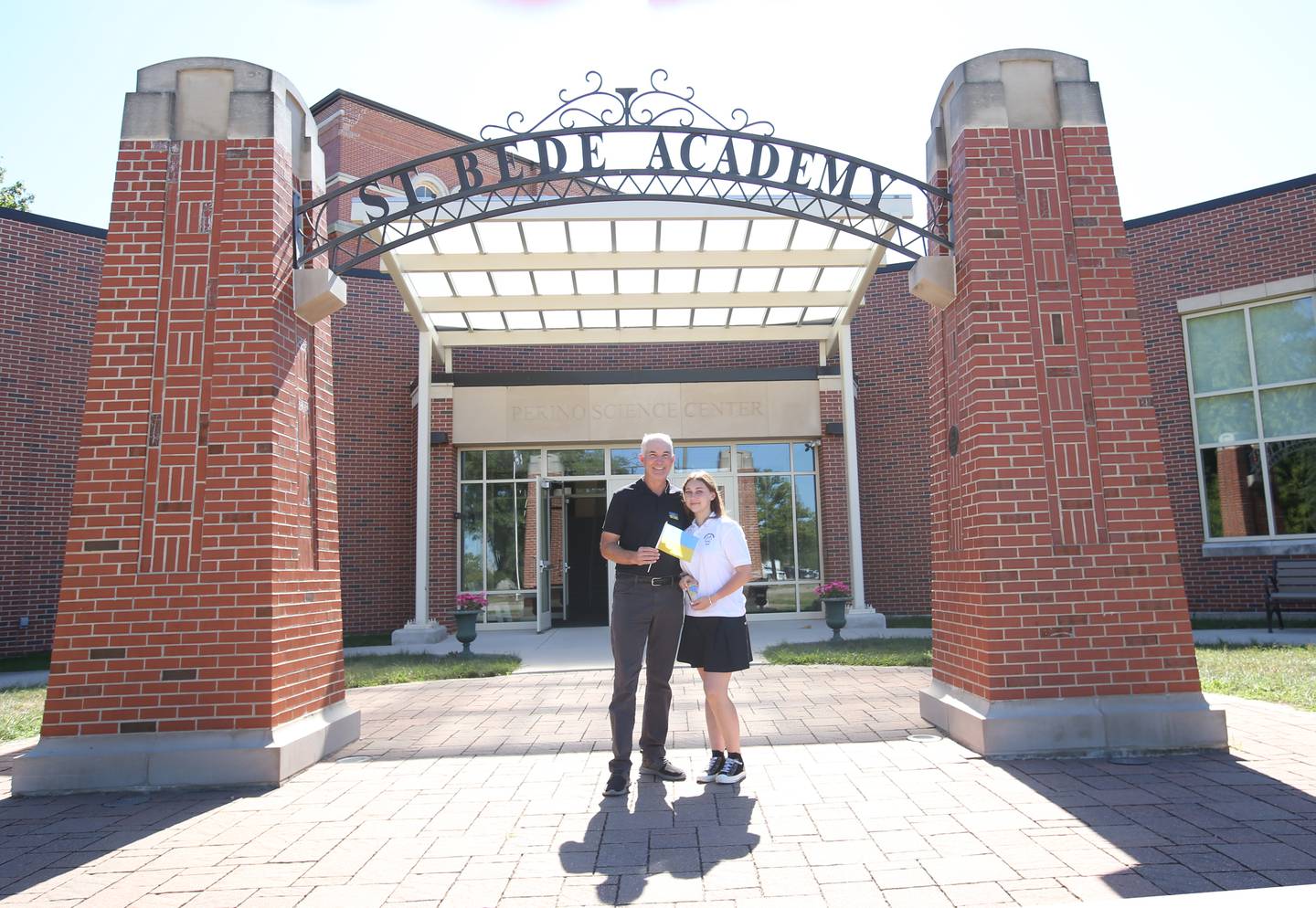 Anna Rudemko a refugee from Ukraine poses with Jim Gamache St. Bede alumnus while holding the Ukraine flag under the arch on Thursday, Aug. 31, 2023 at St. Bede Academy. Rudemko is a student at St. Bede who wanted to come to a school that she could feel welcome and exceed while her country is at war with Russia.