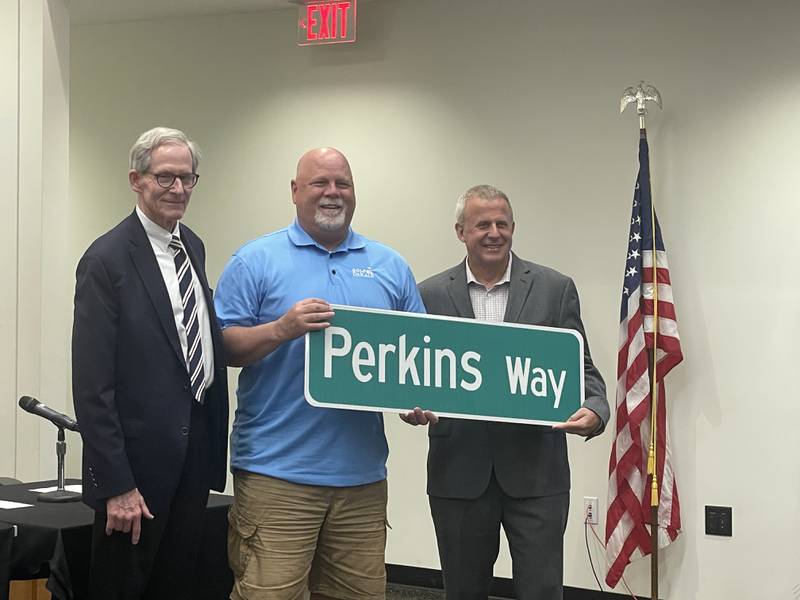 DeKalb City Manager Bill Nicklas, outgoing DeKalb Ward 4 Alderman Greg Perkins and DeKalb Mayor Cohen Barnes (left to right) hoist an honorary street sign May 12, 2025, during a DeKalb City Council meeting.