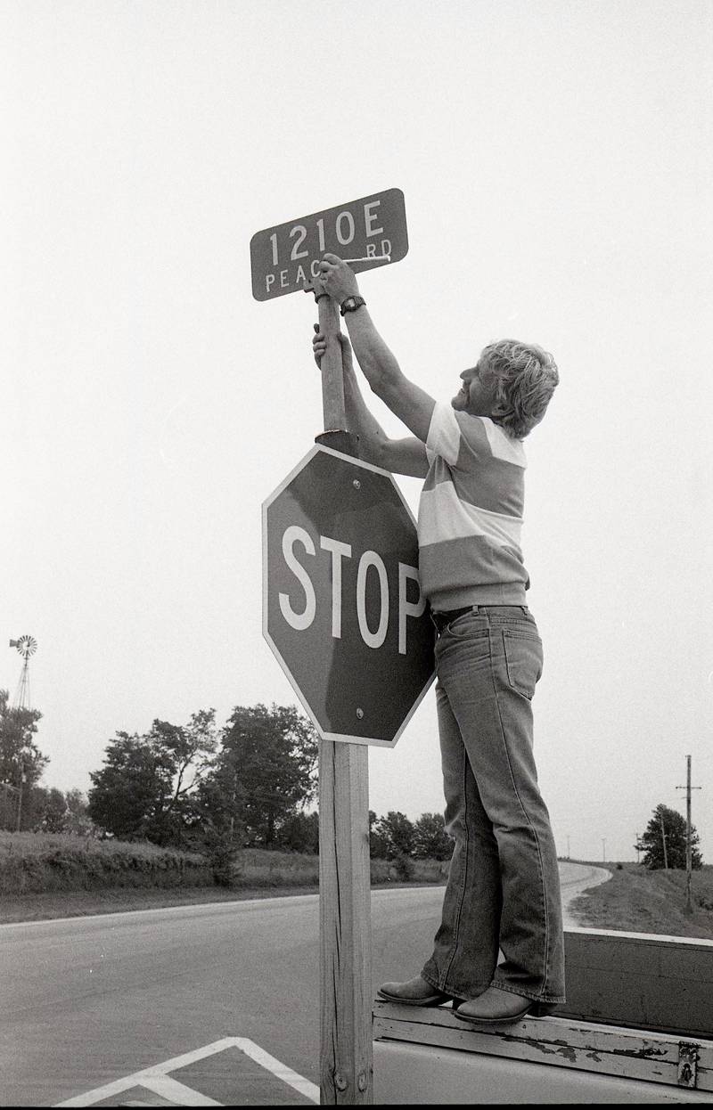 Peace Road, named by eight-year-old Malta student Catherine Rogers, was dedicated with a ribbon-cutting ceremony on August 22, 1988. Rick Buehler of DeKalb, Traffic Control Technician for the Dekalb County Highway Department, installs a sign with the road's new name at the intersection with Barber Greene Road.