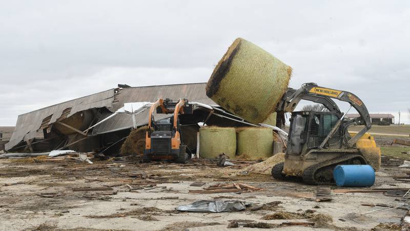 Rob Gunderson uses a skid steer to move a hay bale from one of the damaged storm sheds as a friend holds the shed's roof on Saturday. Friday night's storm ripped through his dairy farm west between Shannon and Forreston. No livestock were injured.