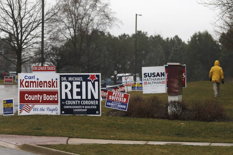 A woman walks past campaign signs that line the driveway to the McHenry County Administration Building, an early voting site, on Friday, March 8, 2024, in Woodstock.