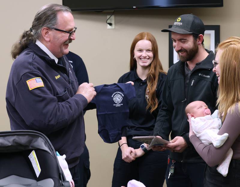 Genoa-Kingston Fire Department Chief Bruce Kozlowski presents Eleanor Lee Altepeter-Knotts, and her parents Brodie Knotts and Sammie Altepeter (right) from Kingston with a Genoa-Kingston Fire Department onesie Thursday, Nov. 2, 2023, as the baby and her family visit the Genoa-Kingston Fire Department in Genoa. An ambulance crew from the department delivered the baby in the ambulance along the side of the road Sept. 29 when they realized they weren’t going to make it to the hospital in time.