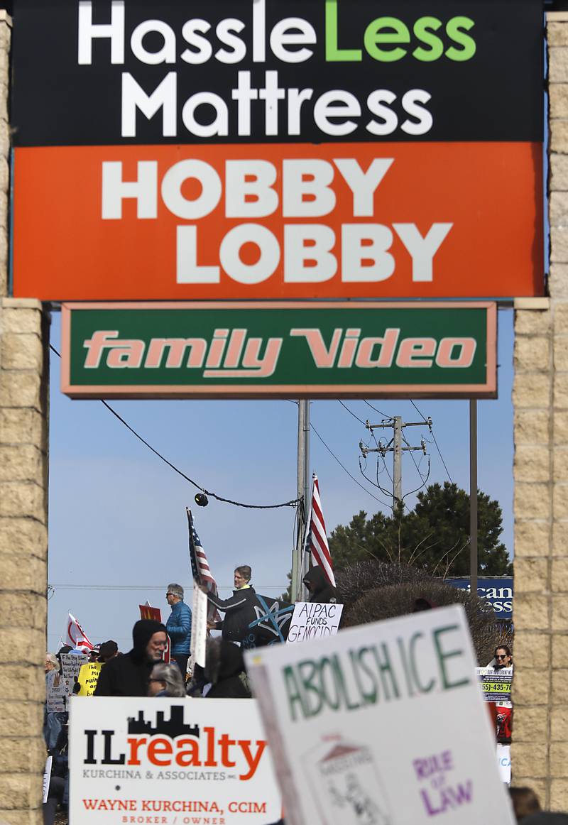 Protesters are framed by a retail signs as they line State Route 31 near the intersection of McCullom Lake Road in McHenry to protest their discontent with President Donald Trump and his administration's policies on Saturday, March 28, 2026, during the McHenry County No Kings Protest. According to an organizer, over 4,000, people took part in the protest.