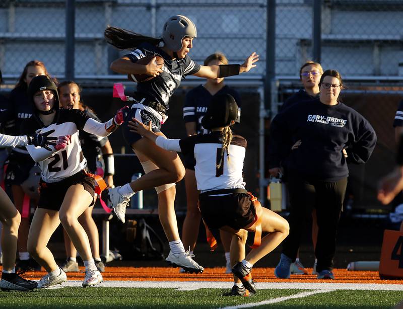 Cary-Grove's Kennedy Manning (center) leaps as she runs along the sideline between McHenry's Lyla Oeffling (left) and Brianna Amedio (right)  during the IHSA McHenry Regional girls flag football championship game on Wednesday, Oct. 8, 2025, at McCracken Field in McHenry.