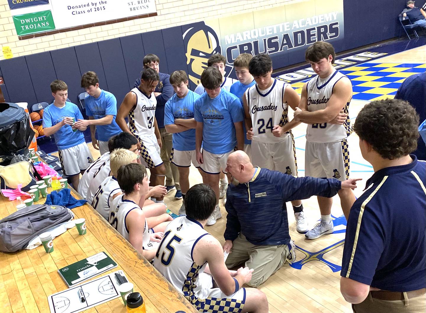 Marquette Academy boys basketball coach Todd Hopkins (kneeling) gestures to the court while talking with his Crusaders during a timeout Tuesday, Jan. 7, 2025, at Bader Gym in Ottawa.