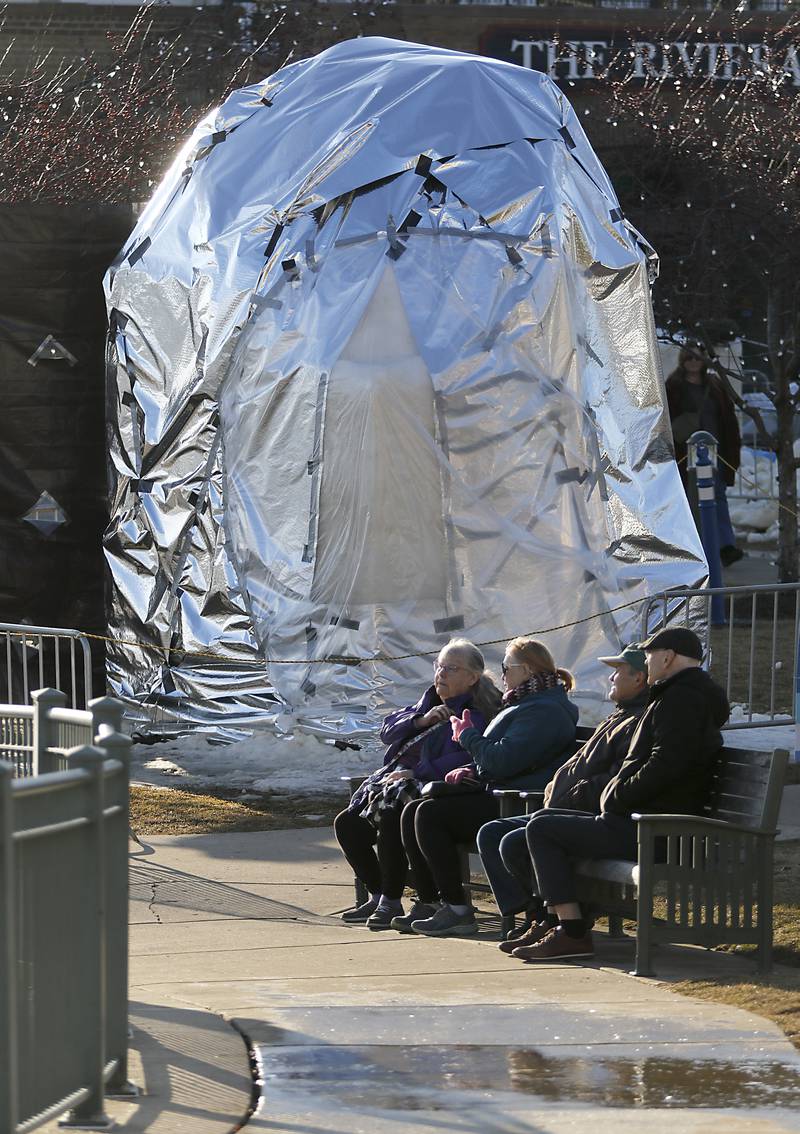 People sit on bench near a covered block of snow to protect it from rain and heat on Thursday., Jan. 30, 2025, Snow carvers have started carving their 15 massive snow sculptures for America’s Snow Sculpting Invitational during Winterfest in Lake Geneva. Winterfest, runs through Feb, 2, 2025. The warm and rainy weather has slowed down the carving of the snow.