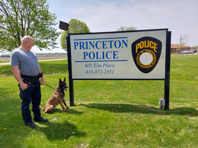 Princeton officer Erik Sorenson watches as K9 officer Gus poses in front of the Princeton Police Department sign.