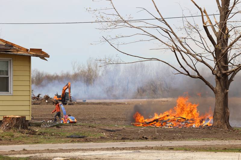 A child retrieves a toy as burn piles are underway behind a home on Strasma North Drive in Aroma Township on April 8, 2026, nearly one month after the EF-3 tornado.