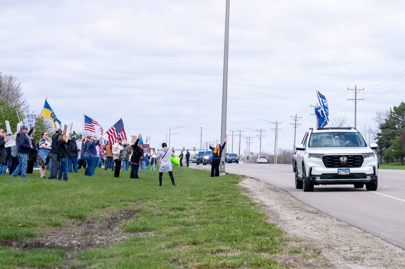 A motorist with a Trump flag displaying outside their vehicle drives past anti-Trump and anti-Musk protesters along Randall Road in South Elgin, IL on Saturday April 19, 2025.  Hundreds of protesters participated in the latest Hands Off protest occurring in the Chicagoland area and the rest of the country.