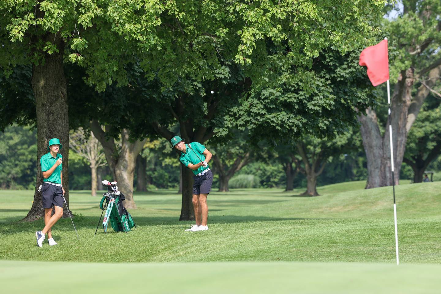 Bishop McNamara's Preston Payne, right, chips toward the green as teammate Griffin Meier looks on during the Bill Breeden All-Area Classic at the Kankakee Elks on Monday, Aug. 18, 2025. Payne scored a 97 as Meier scored a 91 to help earn Bishop McNamara fourth place overall.