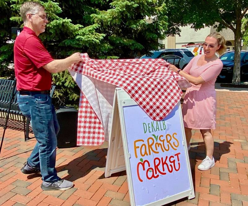 DeKalb Chamber of Commerce executive director Matt Duffy (left) and chamber marketing events manager Virginia Filicetti unveil a new logo for the DeKalb farmers market on the opening day of the market's 30th year on Thursday, June 5, 2025, in Van Buer Plaza in downtown DeKalb.
