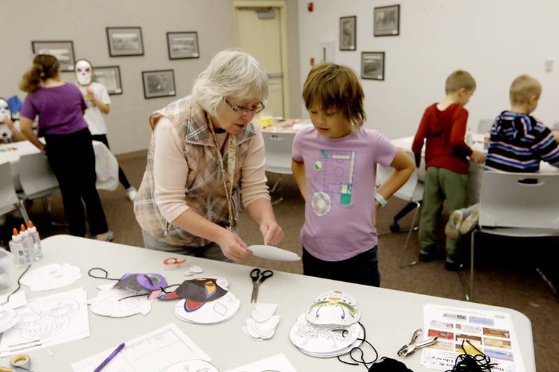Karen Sutera, the Director at Harvard Diggins Public Library, explains to Violet Wargaski, 9, mask options and how to make a mask as children make masks on Wednesday, Oct. 23, 2024, during at Kids Crafternoon at the Harvard Diggins Library. Children were able to chose between several Halloween themed masks to make.