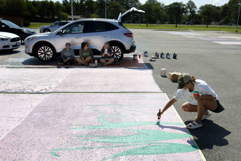 Rubi Gonzalez, Gabriela Gonzalez and Summer Hertel take a break in the shade of a car as painter Payton Walsh paints tulips on butterflies on Hertal’s parking spot on Tuesday, Aug. 13, 2024, at Marian Central High School in Woodstock.