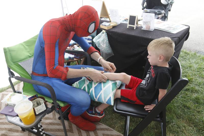 Spiderman, played by Vaughan McMillan, applies a glitter Spiderman tattoo on Landon Harvey’s leg Friday, July 1, 2022, during Lakeside Festival at the Dole and Lakeside Arts Park, 401 Country Club Road in Crystal Lake. The festival continues noon to 11 p.m. July 2 and noon to 10 p.m. July 3. The festival features bands on two outdoor stages, food and drinks, a baggo tournament, and carnival rides and games. Among the activities for kids are face painting, a balloon twister, a stilt walker, team mascots and a magician.