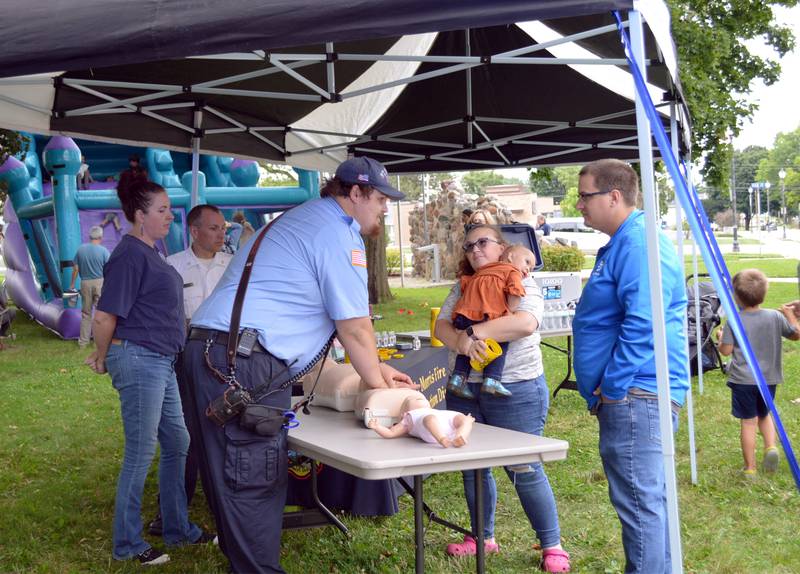 Rachel Gabrielson, of Mt. Morris, (center) holds 20-month-old daughter Elizabeth while she and husband Steven Gabrielson (right) listen and watch Mt. Morris firefighter and paramedic Frank Harenza explain and demonstrate proper CPR on children on Tuesday, Aug. 6, 2024, during the village's first National Night Out.