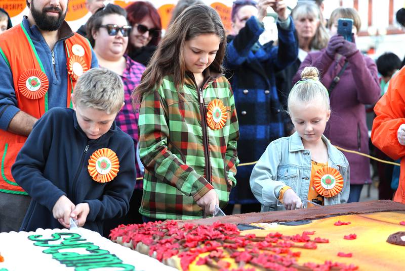 Mason Clark (left) honorable mention in the theme contest, Vivian Rubicz, theme contest winner, and Lennox Freeman, (right) honorable mention, take the first cut out of the giant cakes donated by Hy-Vee during the cake cutting ceremony Wednesday, Oct.26, 2022 on North Maple Street near the DeKalb County Courthouse during the first day of the Sycamore Pumpkin Festival.