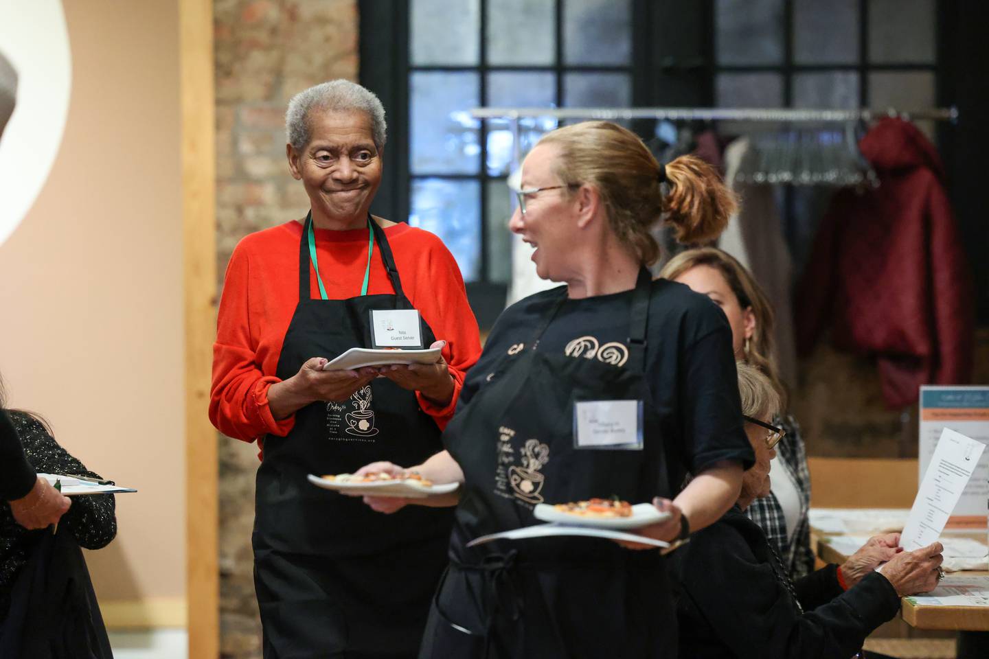 Guest server Nita Moffett, left, smiles as she carries a food order to a table with  helper Tiffany Heneghan during a Mistaken Orders program event, hosted by nonprofit Our Aging Services and MCA Senior Adult Day Center at Stefari West Avenue on Nov. 18, 2025. Heneghan is a board member with MCA Senior Adult Day Center.