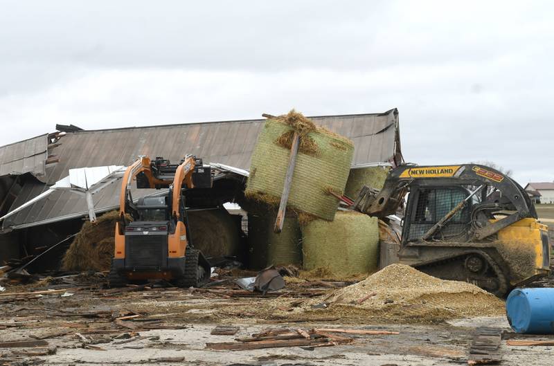 Rob Gunderson uses a skid steer to move a hay bale from one of the damaged storm sheds as a friend holds the shed's roof on Saturday. Friday night's storm ripped through his dairy farm west between Shannon and Forreston. No livestock were injured.
