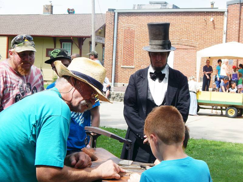 Abraham Lincoln impersonator Max Daniels, in character as Lincoln, watches Gary Eggleston explain to Tristan Melton of Sycamore how to operate a pedal-powered scroll saw at the Kishwaukee Valley Heritage Society's annual Pioneer Day.