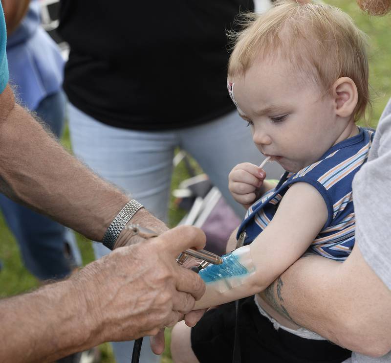 Grayson Graham calmly enjoys his candy sucker while an airbrushing tattoo flag is applied to his arm Tuesday, Aug. 8, 2023, during Unlimited Fun Day at City Park in Streator. The event was sponsored by Streator Unlimited.