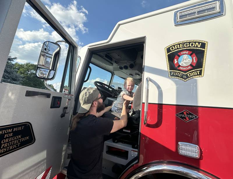 Ezra Towle, 22 months, gets behind the steering wheel of the Oregon Fire Department's ladder truck with the help of his dad, Eric,  while checking out the "Touch-A-Truck" event held during the River's Edge Farmers Market in Oregon on Thursday, June 26, 2025.