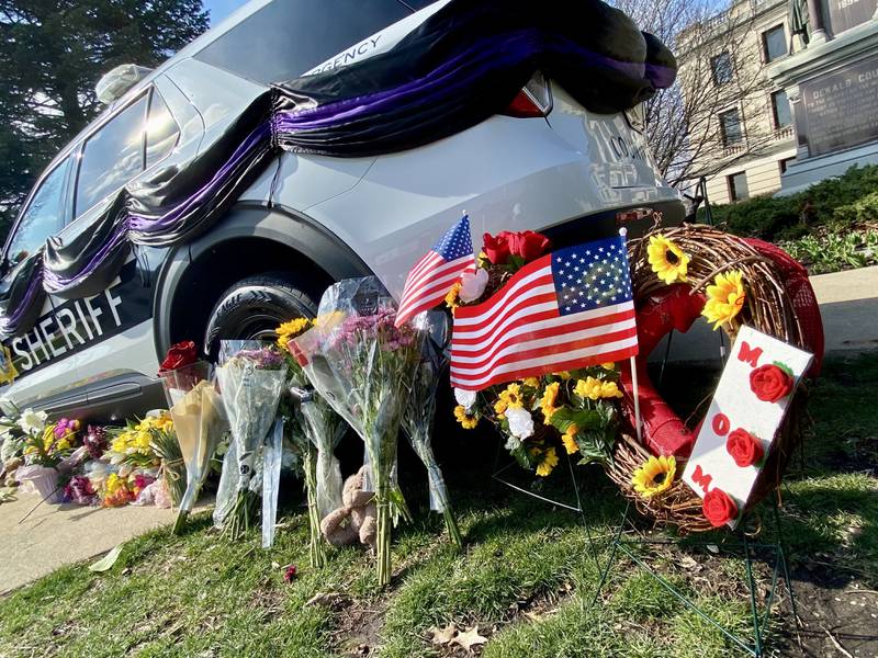 A floral wreath with the word "Mom" on it stands next to a DeKalb County Sheriff's Office vehicle displayed on the DeKalb County Courthouse lawn in Sycamore on Saturday, March 30, 2024. Mourners have visited the site to leave trinkets, flowers and other tokens in memory of sheriff's deputy Christina Musil, who was killed while on duty after her squad car was rear-ended by a truck Thursday, March 28, 2024.