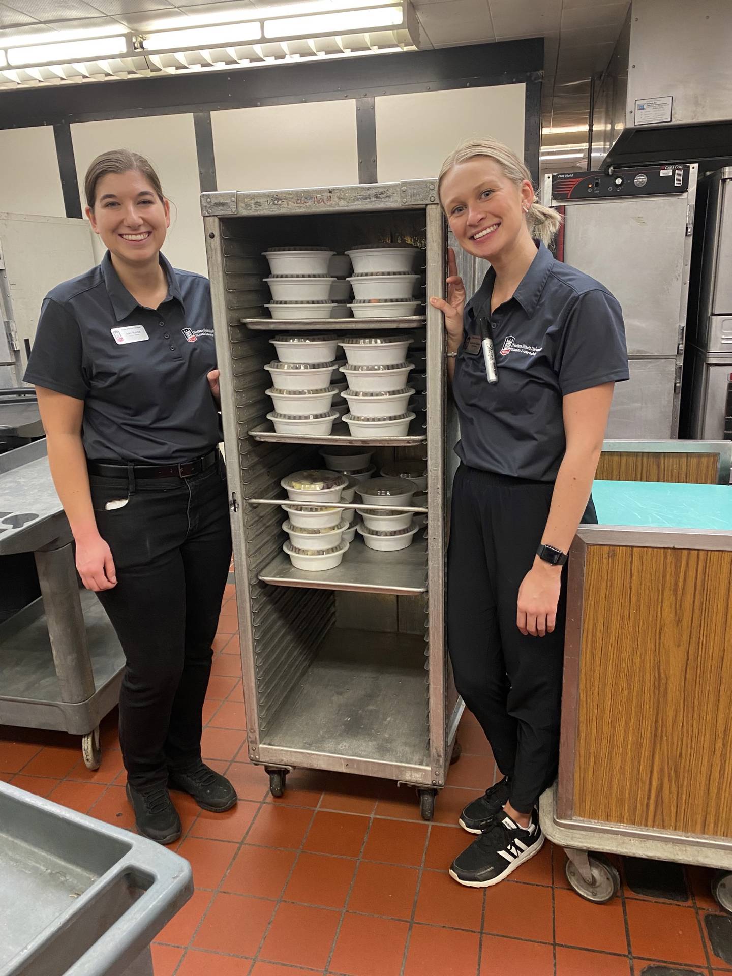 Dietetic interns Amy Martin and Gabby Glesner stand next to food packaged as a part of the Huskie Harvest program, which recovers food destined to be wasted and repackages it into a free meal for food insecure students. Photo provided by Northern Illinois University.