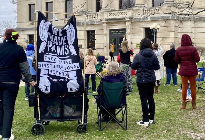 A sign that says, "We wave palms. They waive constitutional rights," is posted next to worship service goers during a Palm Sunday Faith Action event on Sunday, March 29, 2026, in front of the DeKalb County Courthouse in Sycamore. Area Christian ministers organized the event to combat the rise of Christian nationalism in the U.S.