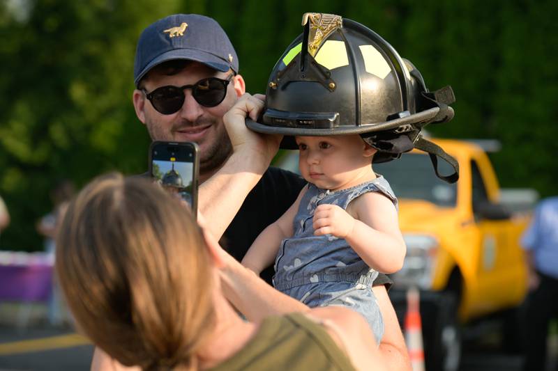 Adam Musial holds his daughter Izabella, 1 as her mom Alex photographs her in wearing a firefighter helmet during Geneva's first National Night Out event on Tuesday, Aug 5, 2025 in Geneva.