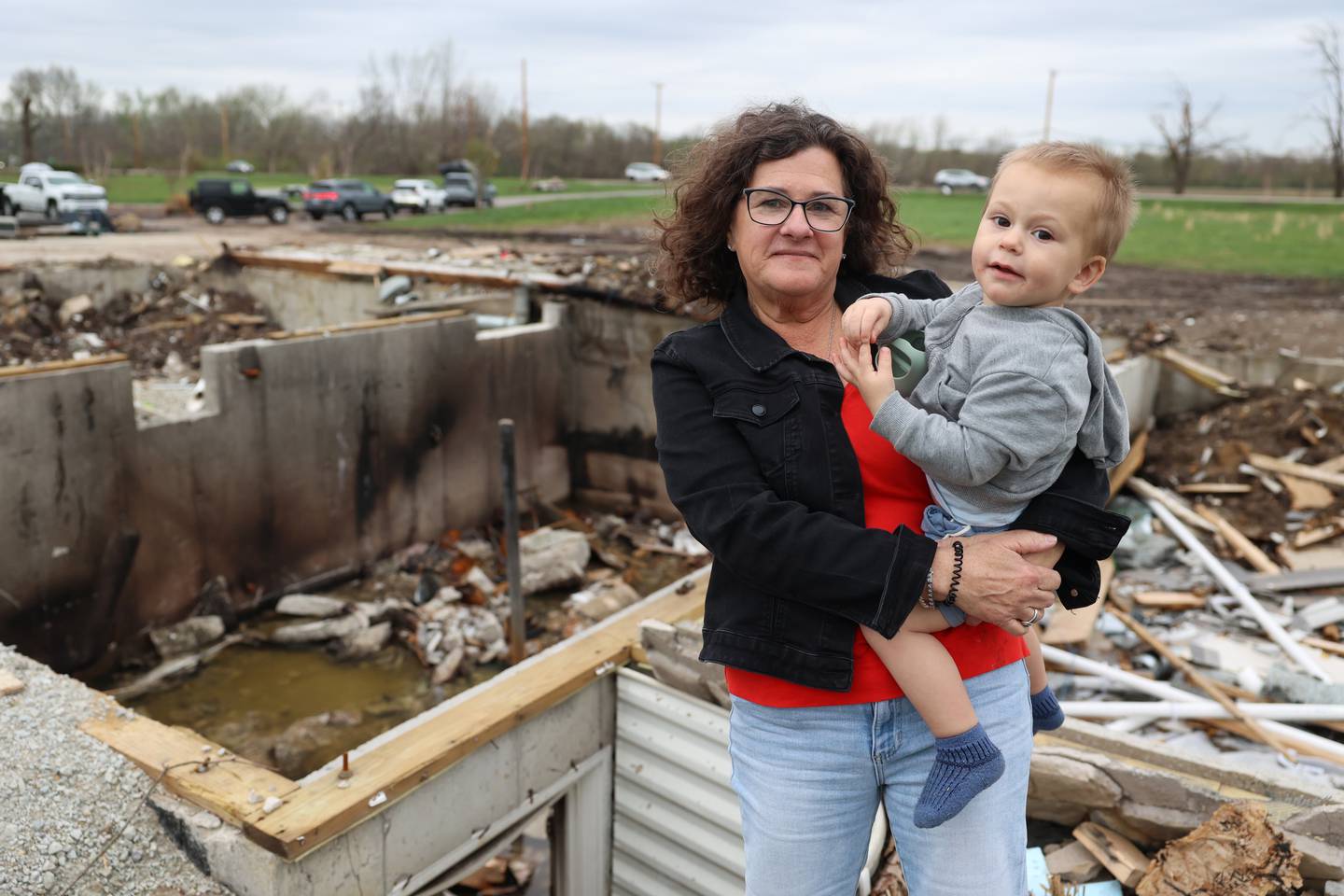 Lisa Gerth holds her grandson Everett, almost 2, on April 15, 2026, outside the basement where she sheltered with family, friends and pets during the March 10 tornado that hit Aroma Township. The Gerth's home on Waldron Road was leveled in the storm, requiring the seven people and four pets to be rescued through basement's window well next to where Lisa stands.