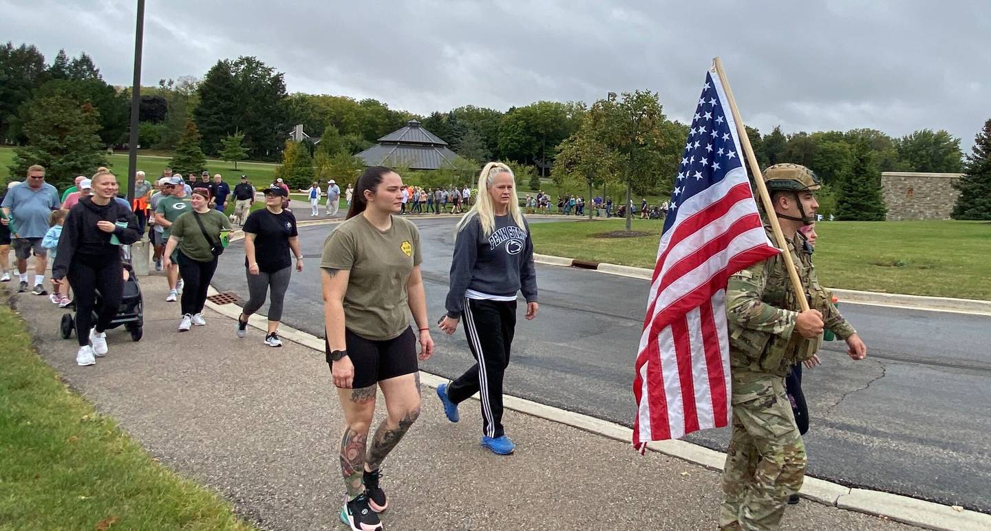 (Right) U.S. Army veteran and reserve member Stephen Cox, 24, of DeKalb, wears his full military garb and hoists an American flag as he joins the line of participants in the inaugural Be the One Walk to raise awareness about veteran suicide rates in DeKalb on Sunday, Sept. 22, 2024. Be the One is part of a national campaign by the American Legion, meant to teach communities how to support veterans' mental wellness.