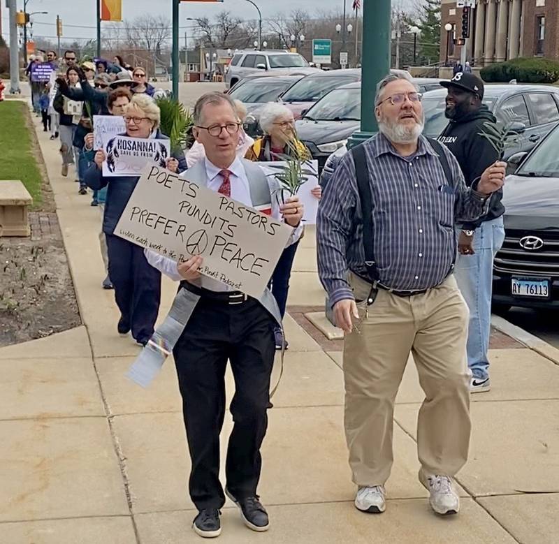 The Rev. John Dorhauer (left) of the United Church of Christ holds a sign and walks with congregants while the Rev. Joe Mitchell of New Hope Missionary Baptist Church (right) directs the procession during a Palm Sunday Faith Action event on Sunday, March 29, 2026, in front of the DeKalb County Courthouse in Sycamore.