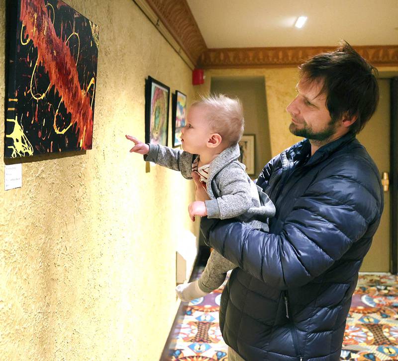 Luke Bujarski, from Sycamore, and his 7-month-old son Aleks, look at a piece by Isabelle Smith, 14, from DeKalb, during the Spring Art Exhibit opening reception Tuesday, March 22, 2022, at the Egyptian Theatre in DeKalb. The exhibit, featuring work from DeKalb County artists, runs until April 25.