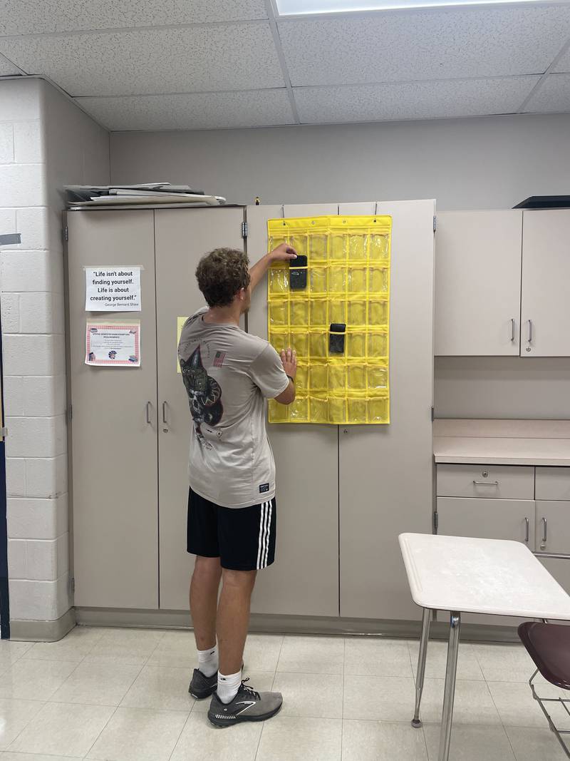 Mendota High School student Travis Mumm demonstrates how to use the cellphone holder in a classroom Wednesday, Aug. 14, 2024. Students place their phones in these holders during class time.