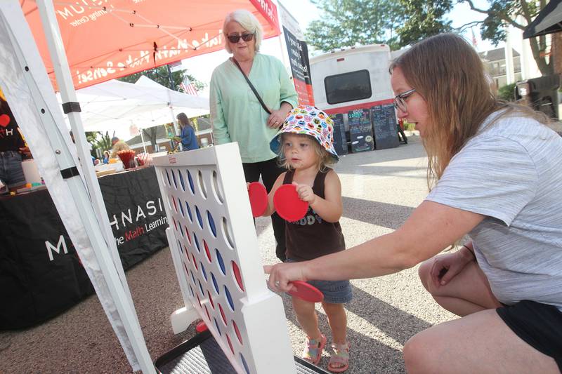 Grace Ayer, 2, of Newport News, Va., (center) plays Connect 4 with her nana, Kim Lettau, of Wauconda and mother, Casie, at the Wauconda Farmers’ Market in downtown Wauconda.  The game was featured at the Mathnasium booth. The farmers’ market runs on Thursday afternoons from 4-7pm through September 29th.