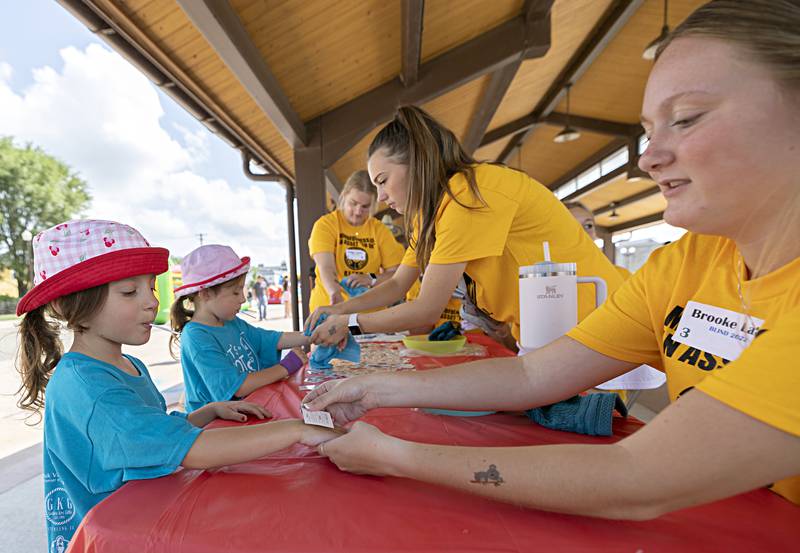 Twins Amelia (left) and Koralina Diedrich, 6 of Sterling, get some new temporary ink by BLIND leaders Izzy Allen (left) of Newman and Brooke Lalley of Prophetstown Friday, August 4, 2023. The group was helping run a family fun fair at the Sterling Market Place as part of Hot Dog Days.