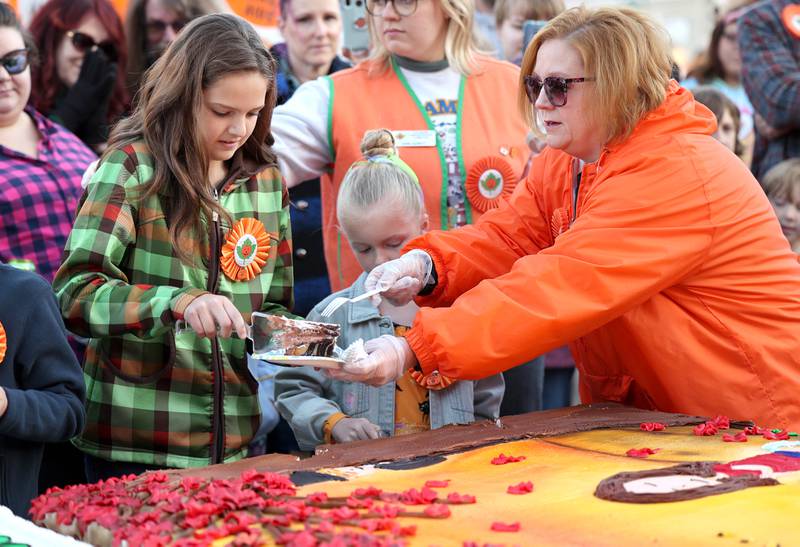 Vivian Rubicz, theme contest winner, gets a hand Jennifer Diehl, from the Sycamore Pumpkin Festival Committee, as she serves up the first piece of the giant cakes donated by Hy-Vee during the cake cutting ceremony Wednesday, Oct.26, 2022 on North Maple Street near the DeKalb County Courthouse during the first day of the Sycamore Pumpkin Festival.