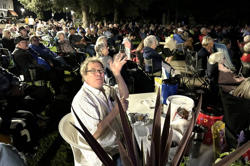 Jamboree Concert Series organizer, Larry Ubben, of Mt. Morris, waves as he eats a burger while seated in the middle of an estimated crowd of 2,500 who turned out to listen to Fortunate Sons, a Creedence Clearwater tribute band, on Friday, Aug, 29, 2025 in Mt. Morris.