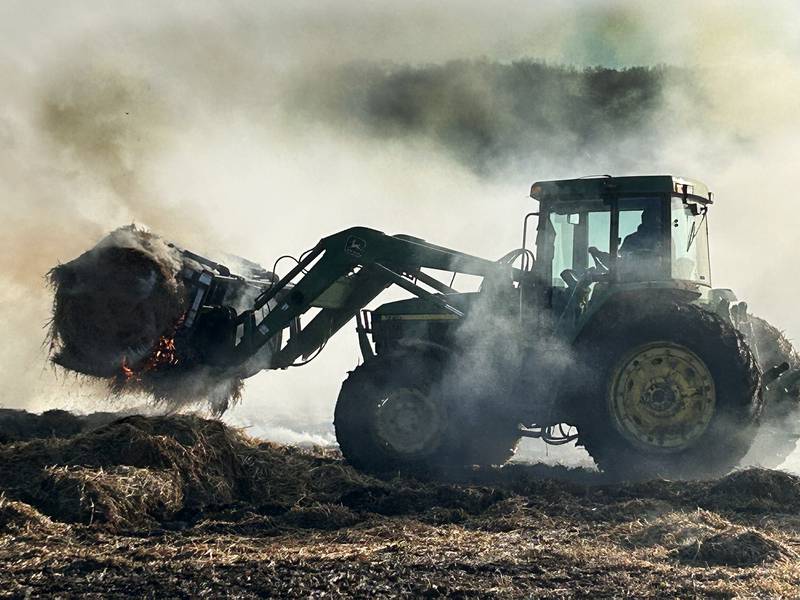 Firefighters from several departments responded to a field and hay bale fire at a farm on West Pines Road between Polo and Oregon on Monday, Feb. 23, 2026. Here, a farmer uses a tractor to carry one of the burning bales out to an open area where firefighters could extinguish it.